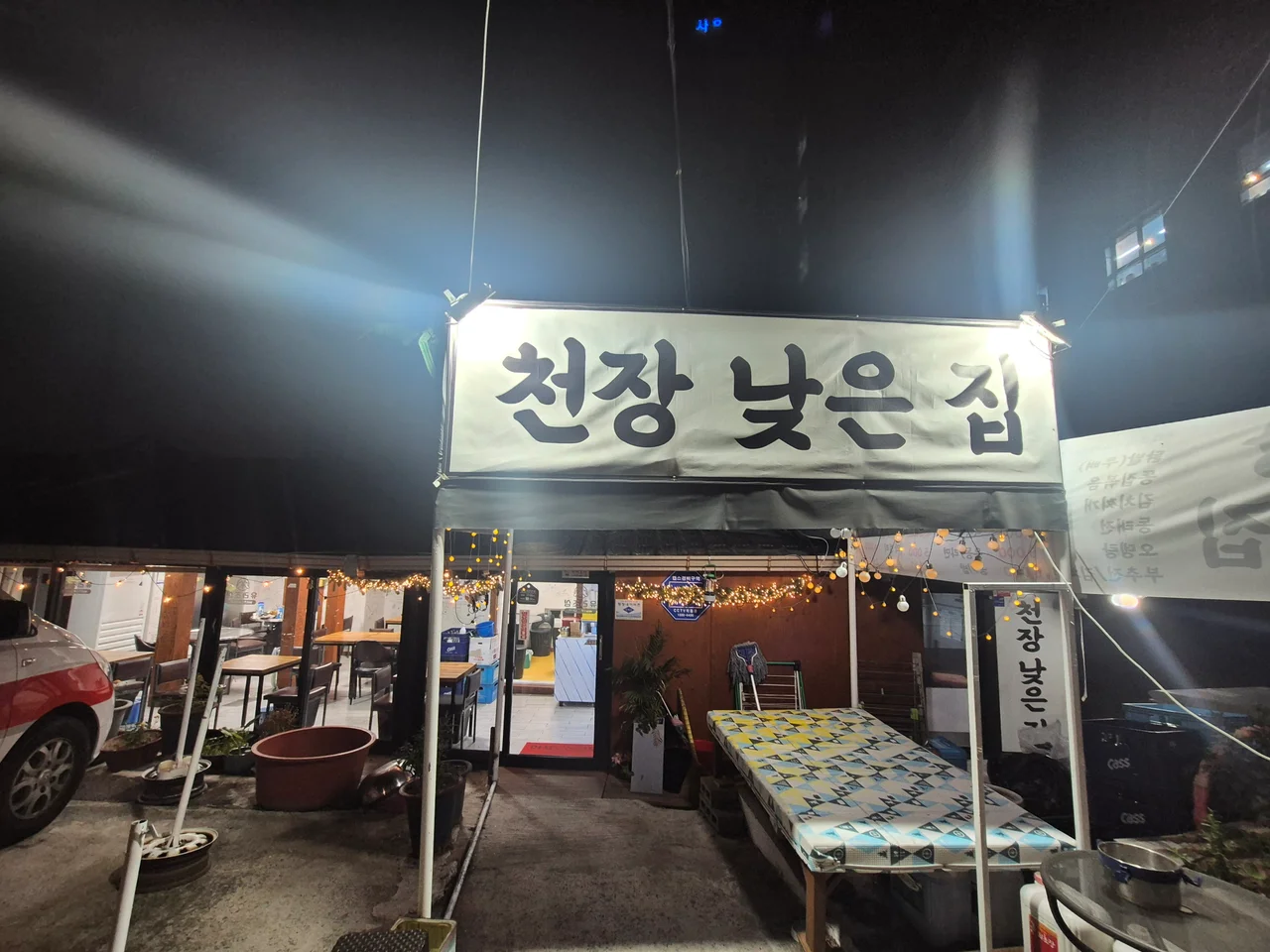 따뜻한 전구 조명과 한글 간판이 걸려 있는 천장낮은집 야외 테라스 입구
