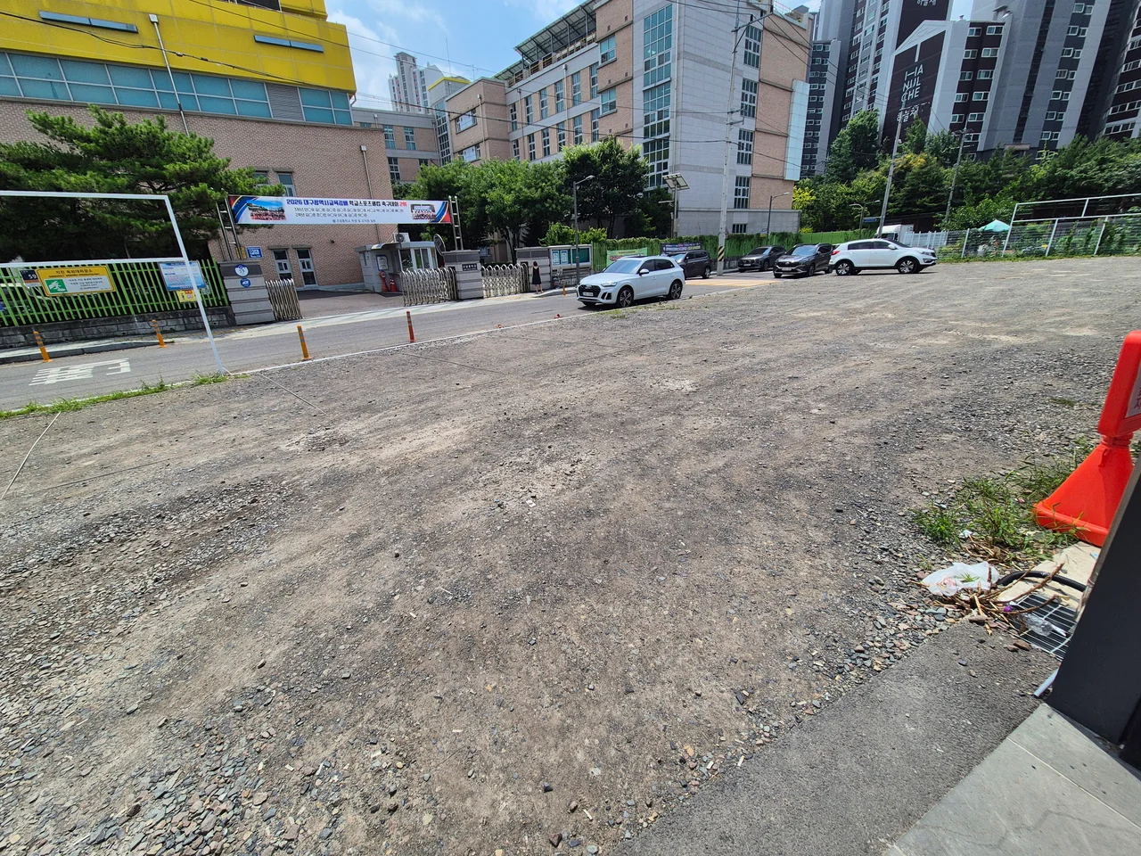 인근 학교 정문과 푸른 나무들이 어우러진 넓은 야외 주차장 풍경