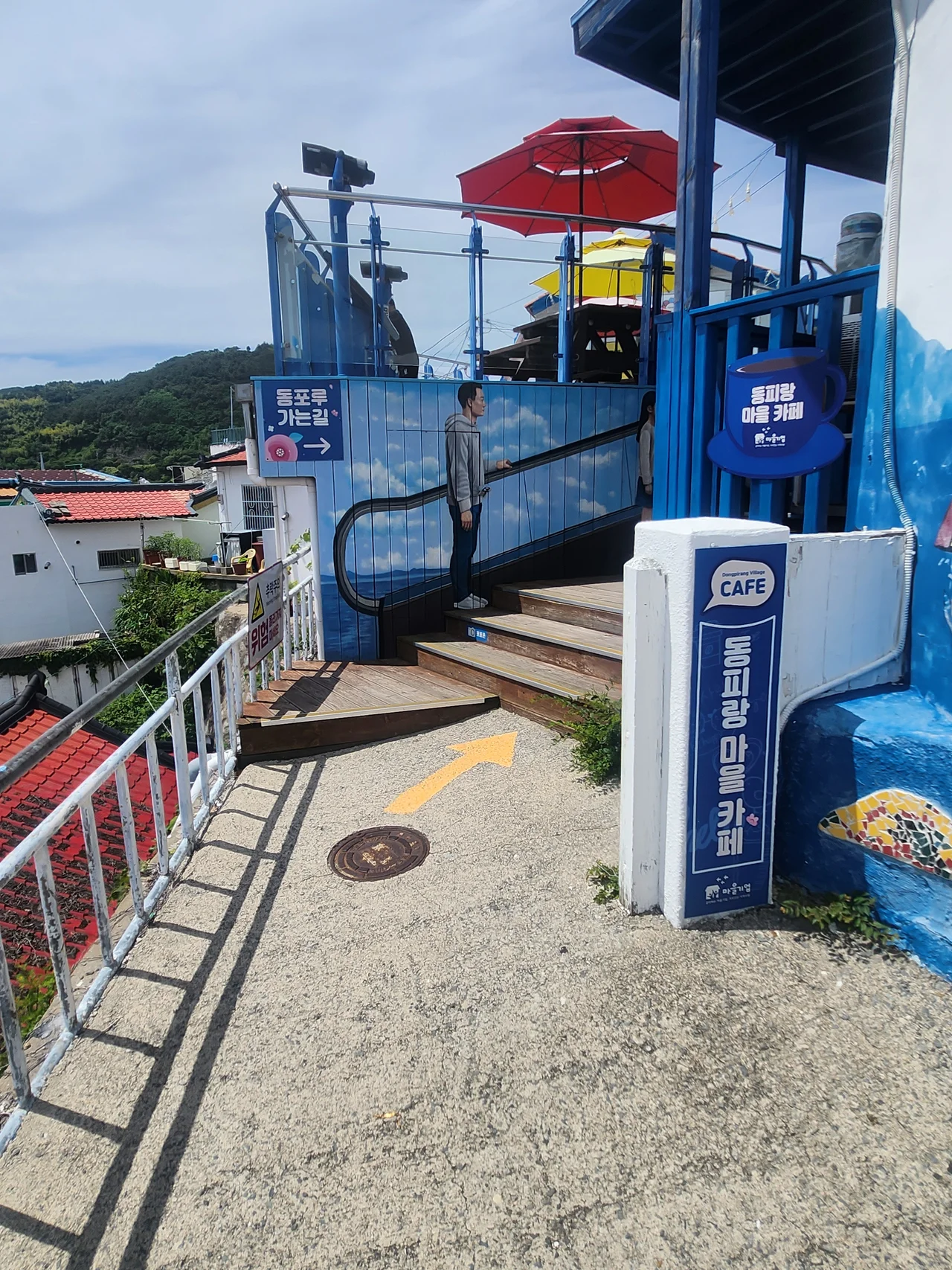 노란 화살표가 그려진 바닥과 하늘 벽화가 그려진 야외 카페 공간